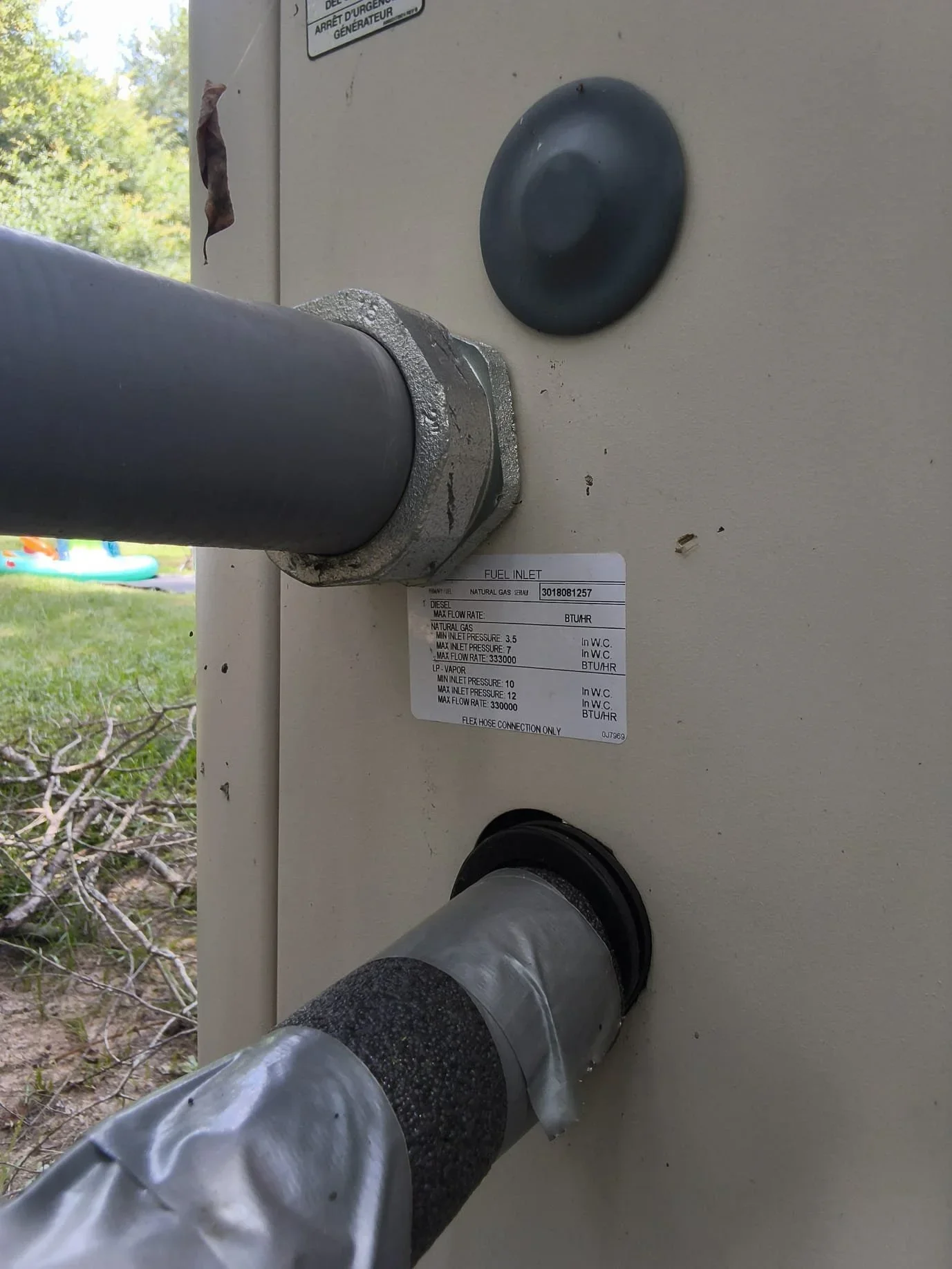 Natural gas fuel inlet with black PVC pipe, compression fittings, and specifications label on generator enclosure