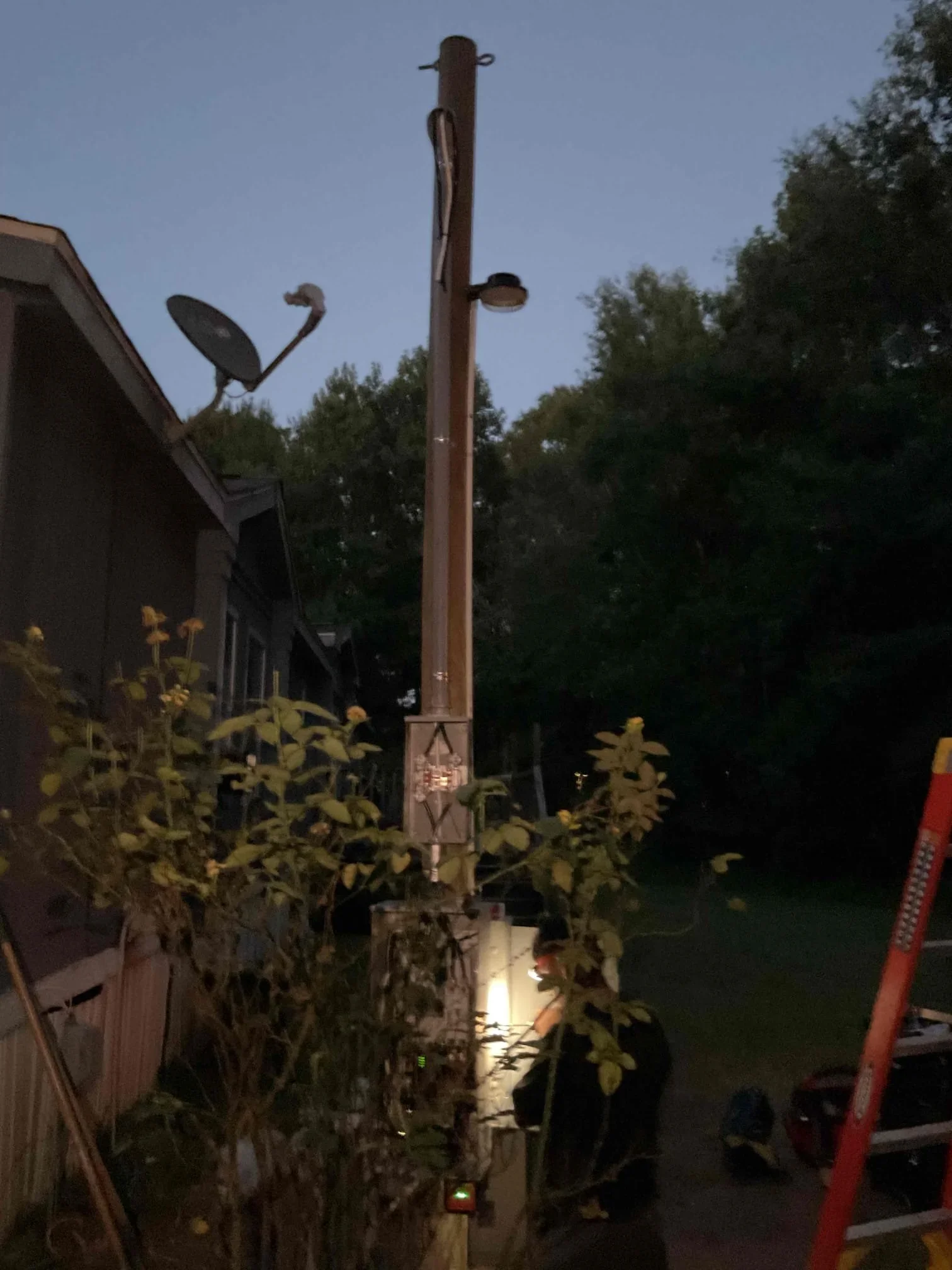 Tall metal utility pole with multiple outdoor light fixtures mounted at different heights, illuminated at dusk