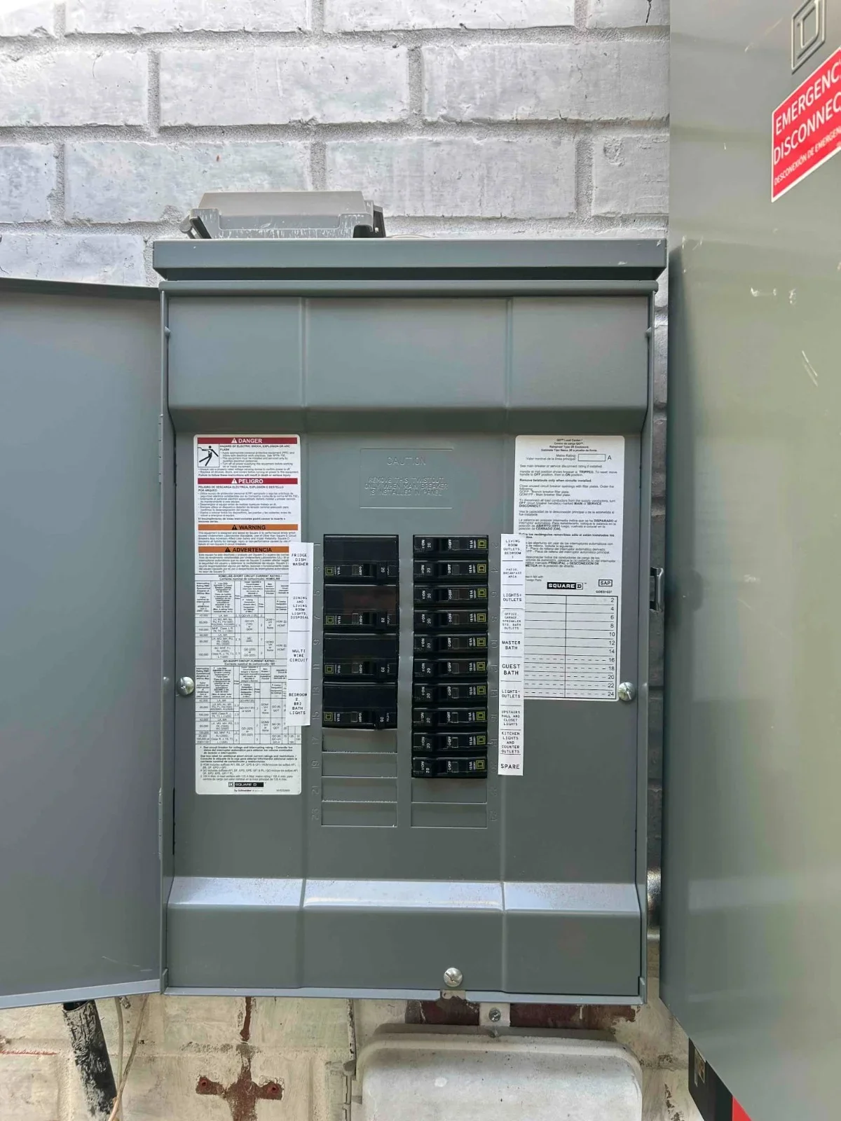 Gray electrical panel with dual-column circuit breakers and safety labels mounted on brick wall